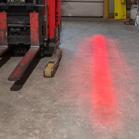 Red fork lift-mounted red illuminated line on concrete warehouse floor.